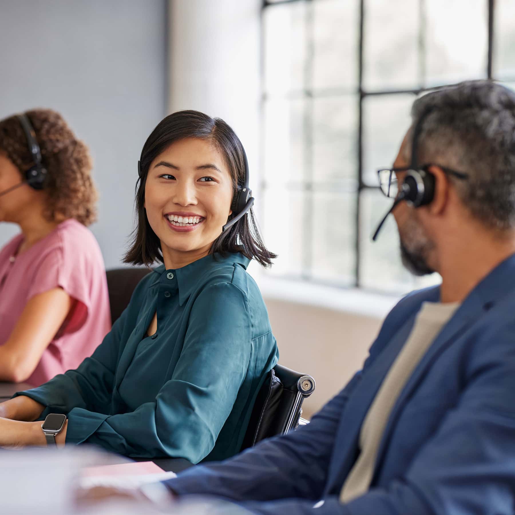 Asian woman in a dark turquoise button up top with telecommunications headset on smiling at colleague