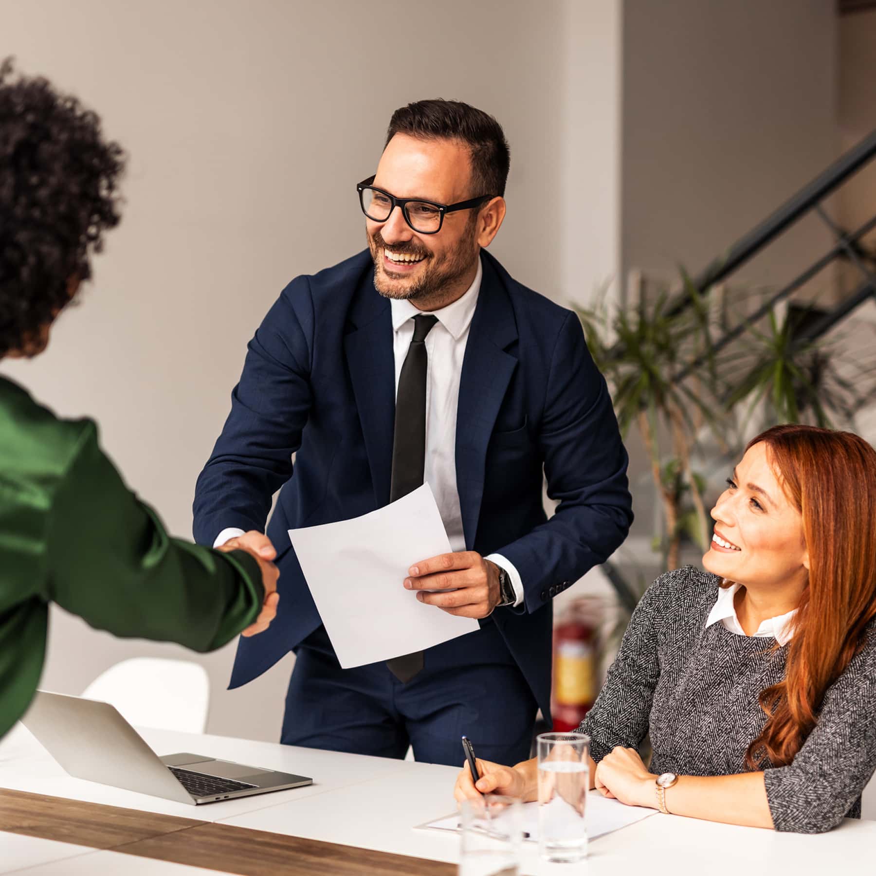 A lady and a man shaking hands in a modern office setting with a happy coworker sitting at table. 